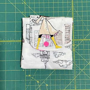 May include: A white fabric pouch with a colorful illustration of a person walking in a city. The illustration includes buildings, streetlights, and clouds. The pouch has a pink and white striped design.