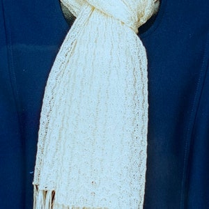 May include: A white knitted scarf with a fringe on the bottom. The scarf is draped over a mannequin wearing a navy blue jacket.