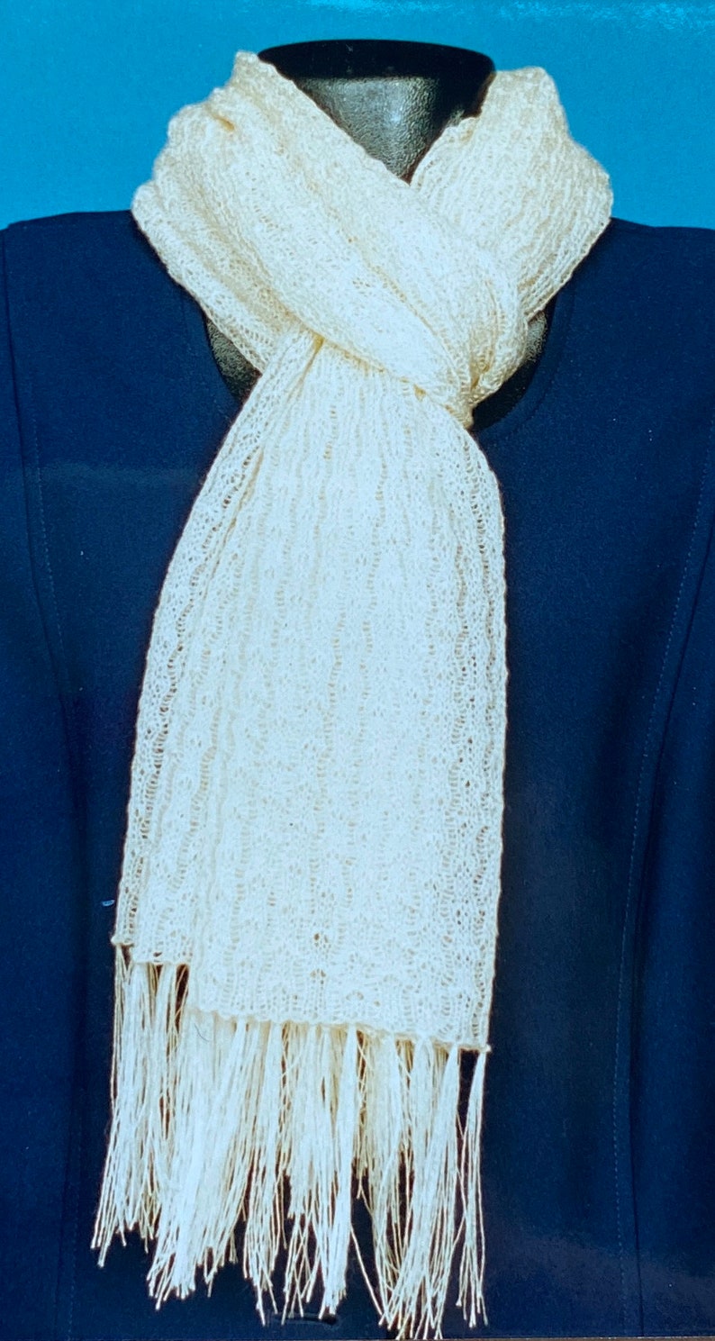 May include: A white knitted scarf with a fringe on the bottom. The scarf is draped over a mannequin wearing a navy blue jacket.