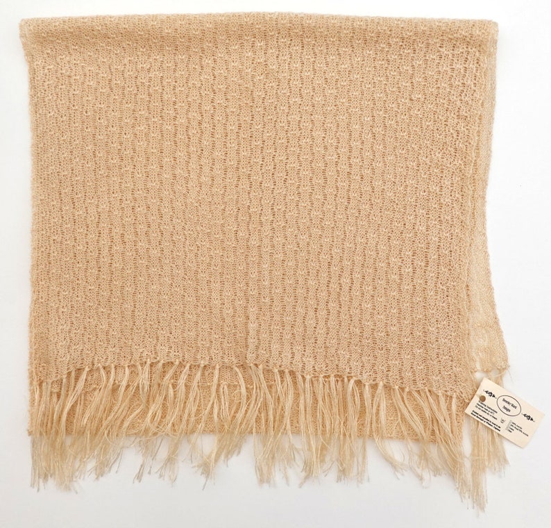 May include: A beige knitted scarf with a fringe border. The scarf is made of a lightweight, open weave fabric. The tag on the scarf reads "Hand Woven Scarf".