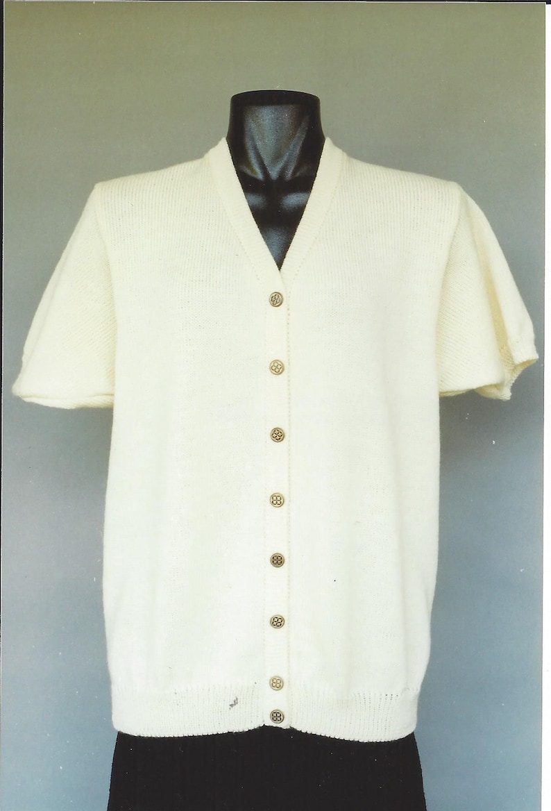 May include: A white short-sleeved cardigan with a v-neck and button closure. The cardigan has a ribbed texture and is made of a soft, lightweight material.
