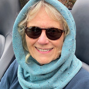 May include: A woman wearing a light blue cowl neck scarf with small holes throughout the fabric. She is wearing round, dark sunglasses and smiling.
