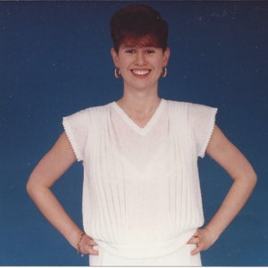 May include: A woman with short, reddish-brown hair is wearing a white, sleeveless blouse with a V-neck. She is smiling and has her arms crossed in front of her. The background is a solid blue.