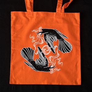May include: Orange tote bag featuring a black and white illustration of two birds in flight. The birds are surrounded by white smoke-like designs.