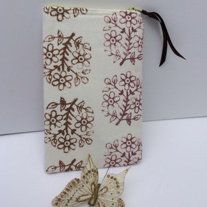 May include: A white zippered pouch with a brown floral print. The pouch has a brown zipper pull and a brown ribbon attached to the zipper. A gold butterfly is in the foreground.