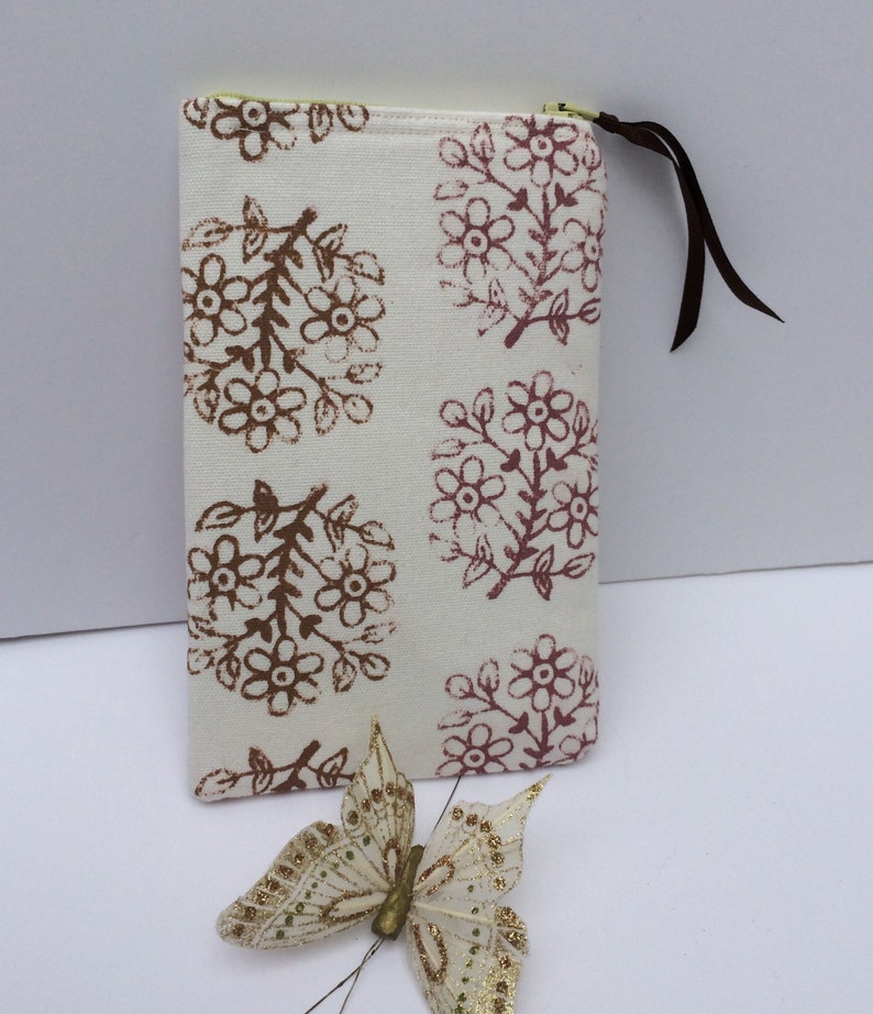 May include: A white zippered pouch with a brown floral print. The pouch has a brown zipper pull and a brown ribbon attached to the zipper. A gold butterfly is in the foreground.