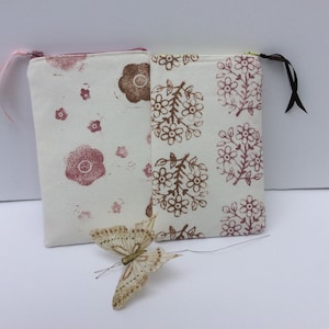 May include: Two fabric pouches with zipper closures. One pouch is white with a pink floral pattern and the other is white with a brown floral pattern. A white butterfly with gold glitter is in the foreground.