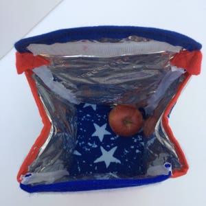 Puede incluir: Una bolsa de almuerzo aislada azul y roja con un forro de patr&oacute;n de estrellas blancas. La bolsa est&aacute; abierta y hay una manzana dentro.