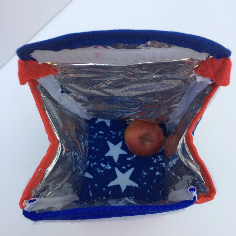 Puede incluir: Una bolsa de almuerzo aislada azul y roja con un forro de patr&oacute;n de estrellas blancas. La bolsa est&aacute; abierta y hay una manzana dentro.