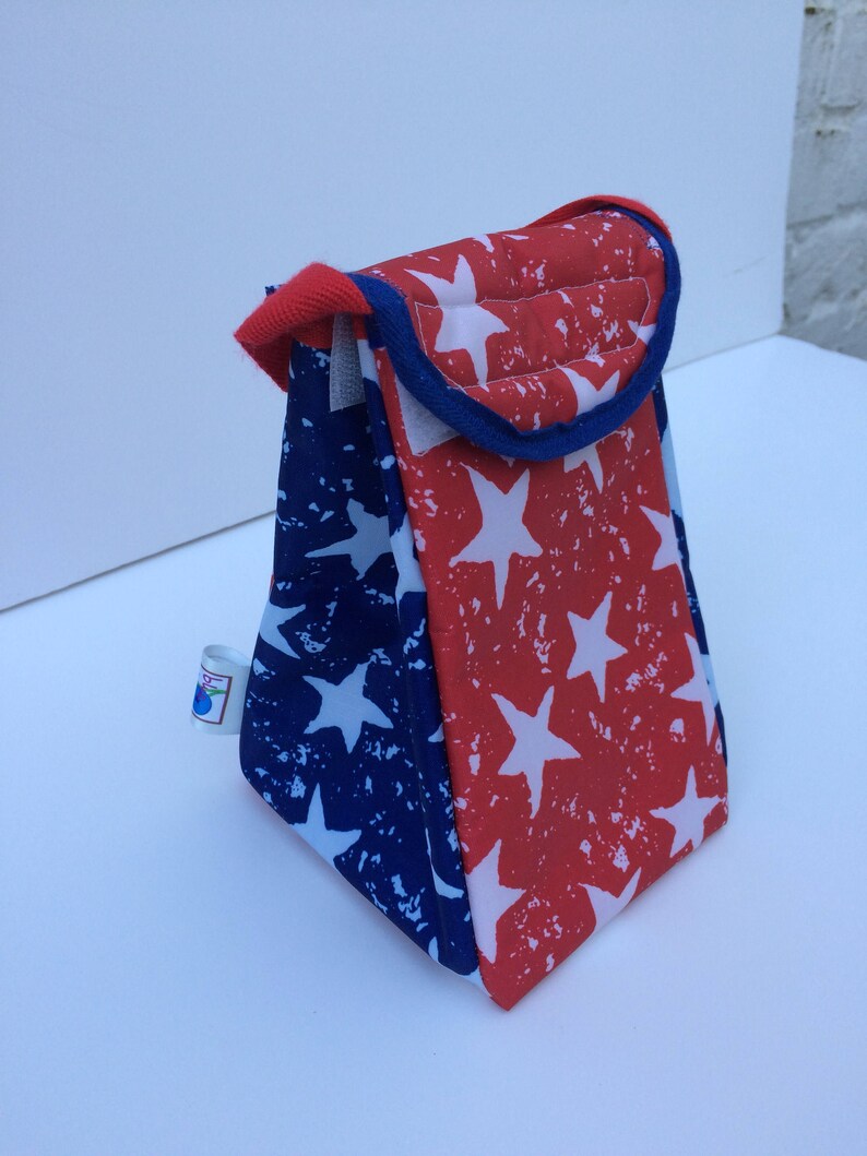 Puede incluir: Una bolsa de almuerzo roja y azul con un patr&oacute;n de estrellas blancas. La bolsa tiene un ribete azul y un asa.