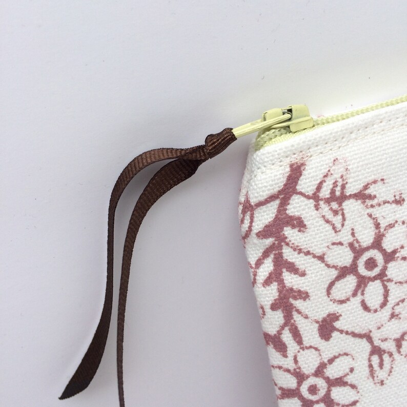 May include: A white canvas pouch with a brown floral print and a brown zipper pull. The zipper pull is attached to a gold zipper.