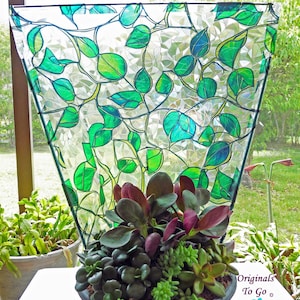May include: A decorative terrarium with a stained glass design featuring green leaves and vines. The terrarium holds a variety of succulents with green and burgundy leaves, in a dark gray ceramic pot. The background shows a sunny outdoor scene.