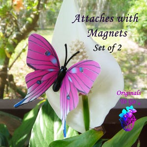 May include: Two pink and blue butterfly magnets with black bodies. The butterflies are attached to a white flower. The text "Attaches with Magnets Set of 2" is visible in the image.