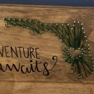 May include: A wooden sign with the text "ADVENTURE awaits" in black lettering. The sign features a string art design of the state of Florida in green thread.
