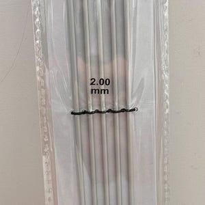 Könnte beinhalten: Packung mit Drops Basic Doppelspitzen-Stricknadeln. Die silberfarbenen Nadeln haben eine Größe von 2,00 mm. Die Nadeln werden von einem schwarzen Band zusammengehalten.