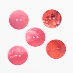 Può includere: Cinque bottoni rotondi in tonalità rosa e rosso sono disposti su uno sfondo bianco. Ogni bottone ha due piccoli fori per cucire. I bottoni variano di colore, dal rosa chiaro al rosso intenso, alcuni mostrando un effetto marmorizzato.