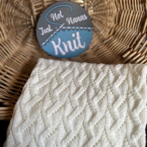 May include: A white knitted blanket with a textured diamond pattern is displayed. The blanket is folded and positioned in front of a round sign with the text "Not Just Havens Knit". The background features a woven basket.