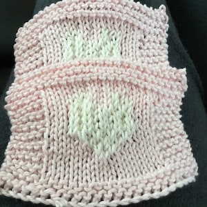 May include: Two knitted squares, one atop the other, in a soft pink hue. Each square features a white heart design in the center. The knitted texture is clearly visible, showcasing the yarn's delicate structure. The squares are likely handmade.