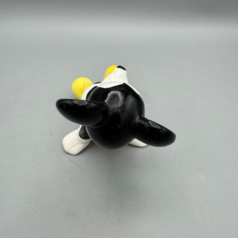 May include: A black and white ceramic figurine of a cartoon dog with yellow ears. The dog is sitting on its hind legs with its front paws outstretched.