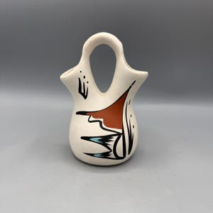 May include: A cream-colored ceramic wedding vase with a central arched handle and two openings. The vessel features hand-painted black, terracotta, and light blue geometric designs on the front, showcasing a smooth matte finish and curved form.