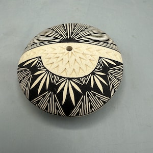 May include: A round, off-white ceramic piece with a central hole. The piece is decorated with black geometric patterns, including radiating lines and stylized leaf shapes. The design is symmetrical and covers the entire surface.