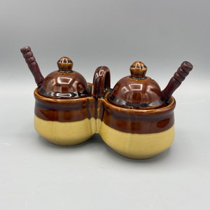 以下が含まれることがあります： 木製のさじが2本付いた、茶色とクリーム色のセラミック製の調味料入れ2個セット。2つの小さな丸い容器には蓋が付いており、大きなベースには持ち手があります。