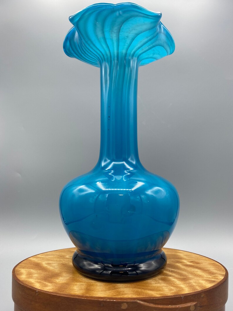 Puede incluir: Un jarr&oacute;n de vidrio azul con una abertura ancha y con volantes y un cuello estrecho. El jarr&oacute;n tiene un patr&oacute;n de espiral y una base negra.