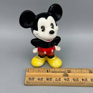 Może przedstawiać: Mała ceramiczna figurka Myszki Miki. Stoi z rękami na biodrach i ma na sobie swoje charakterystyczne czerwone spodenki z jednym ż&oacute;łtym guzikiem i czarne buty. Figurka ma około 5 cm wysokości.