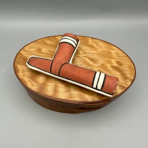 Könnte beinhalten: Eine rote und weiße Keramikpfeife liegt auf einer Holzkiste mit Holzmaserung. Die Pfeife ist L-förmig und hat schwarz-weiße Streifen.
