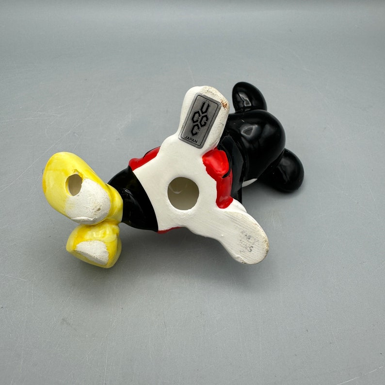 May include: A ceramic figurine of Mickey Mouse in his classic red shorts and yellow shoes. The figurine is made in Japan and has a black and white pattern on the back. The figurine is in a standing position with his arms outstretched.