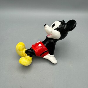 May include: A ceramic figurine of Mickey Mouse lying down. He is wearing his signature red shorts with white polka dots and yellow shoes. Mickey is smiling and looking to the right.