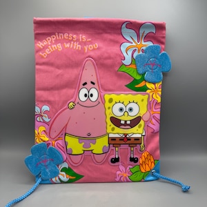 Puede incluir: Bolsa de cordón rosa con los personajes de Bob Esponja y Patricio Estrella. La bolsa tiene un diseño floral y el texto "Happiness is... being with you".