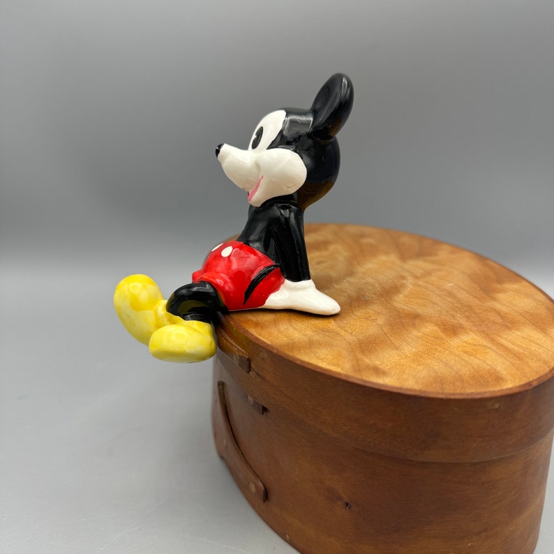May include: A black and white ceramic figurine of Mickey Mouse sitting with his legs crossed. He is wearing red shorts with white polka dots and yellow shoes. The figurine is sitting on a wooden box.
