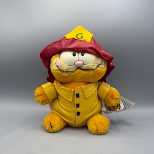 Puede incluir: Un peluche de Garfield disfrazado de bombero. El gato naranja lleva una chaqueta amarilla con botones negros y un sombrero rojo con un emblema amarillo. El juguete tiene una nariz rosa y una expresión sonriente. Una etiqueta está adjunta.
