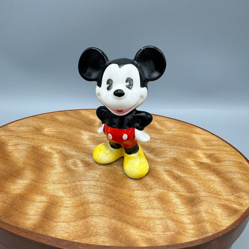 Może przedstawiać: Ceramiczna figurka czarno-biała przedstawiająca Myszki Miki w czerwonych spodenkach i ż&oacute;łtych butach. Figurka stoi na drewnianej powierzchni.