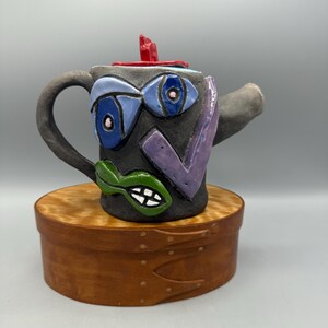 May include: A dark gray ceramic teapot featuring a multi-colored abstract face design with blue eyes, a purple cheek accent, and green lips. The teapot has a red lid handle and a curved spout, showcasing a textured, hand-sculpted appearance.
