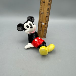 May include: A ceramic figurine of Mickey Mouse in a red and white shorts outfit. Mickey is laying on his side with his left arm extended and his right arm bent at the elbow. He is wearing yellow shoes.