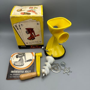 Puede incluir: Un molinillo de alimentos manual de plástico amarillo con base de succión. El molinillo tiene un tazón extraíble y una manivela. La caja incluye un juego de cuchillas de repuesto y un manual. La caja dice "Tritatutto Jolly" y "Jallu".
