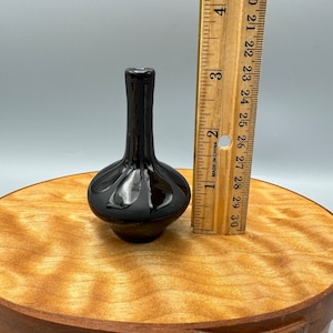 Puede incluir: Un pequeño jarrón de cerámica negro con cuerpo redondeado y cuello estrecho. El jarrón mide aproximadamente 7,6 cm de alto.