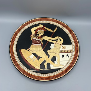 May include: A black and white ceramic plate with a red and gold border. The plate features a scene of a Greek warrior fighting a woman. The warrior is holding a sword and shield, and the woman is holding a spear. The plate is decorated with a black and white geometric pattern.
