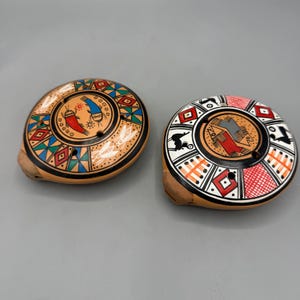 Op de afbeelding: Twee handbeschilderde keramische ocarina's. Eén heeft een ontwerp met rode, blauwe, groene en gele geometrische vormen. De andere heeft een zwart, wit en rood ontwerp met een centrale geometrische figuur en dierensilhouetten. Beide hebben een afgeronde vorm.