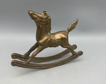 Puede incluir: Una figurita de caballo de balancín de latón de color dorado. El caballo está en posición de galope con la cabeza girada hacia un lado. La base de balancín está hecha de dos piezas de latón curvadas.