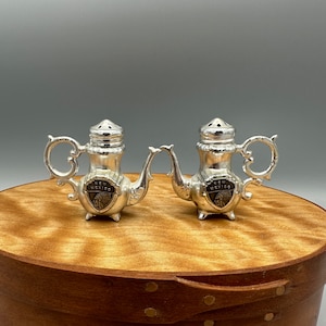 May include: A pair of silver teapot-shaped salt and pepper shakers with a New Mexico state emblem on each. The shakers are on a wooden surface.