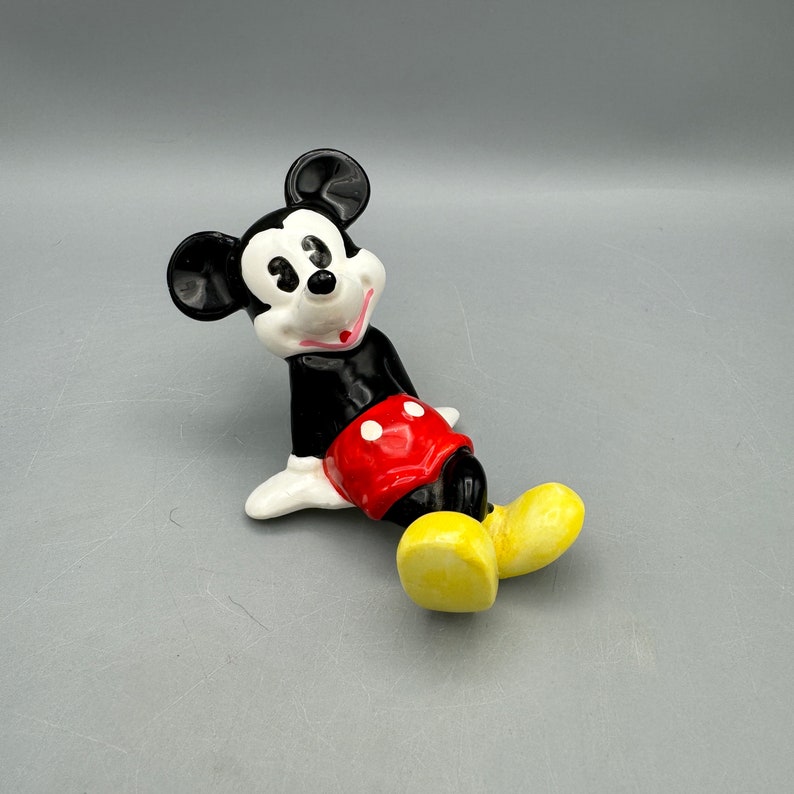 May include: A ceramic figurine of Mickey Mouse in a relaxed pose. He is wearing his signature red shorts with white polka dots, black shoes, and a black and white face with a big smile.