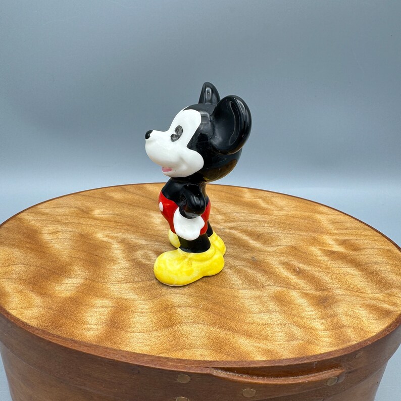 Może przedstawiać: Ceramiczna figurka Myszki Miki stojąca na drewnianej powierzchni. Miki ma na sobie swoje charakterystyczne czerwone spodenki, ż&oacute;łte buty i czarno-białe rękawiczki. Patrzy w prawo.