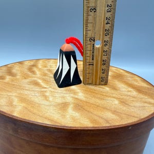 Puede incluir: Un pequeño adorno de madera pintado a mano con una base negra y diseños geométricos blancos. La parte superior es de color madera natural y tiene una cuerda roja para colgar. El adorno mide aproximadamente 2,5 cm de alto.