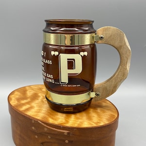 Puede incluir: Una taza de vidrio marrón con un asa de madera. La taza tiene una banda dorada alrededor de la parte superior e inferior. La taza tiene una letra dorada "P" en la parte delantera. La taza está sentada en una caja de madera. El texto en la taza dice "Enco Nat'l Corp 1973".