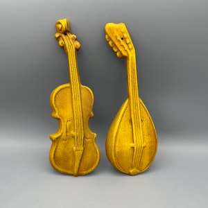 May include: Two gold-coloured musical instruments, a violin and a mandolin, are displayed against a grey background. Both instruments have a detailed, ornate design.