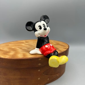 May include: A black and white ceramic figurine of Mickey Mouse sitting on a wooden box. Mickey is wearing red shorts with a white circle and yellow shoes.