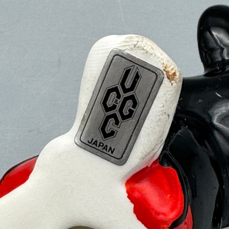 May include: Close-up of a white ceramic figurine with a black and red design. The figurine has a silver label that reads "UCC JAPAN".